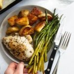A hand holding a small gray plate with a chicken breast, asparagus, and potatoes.