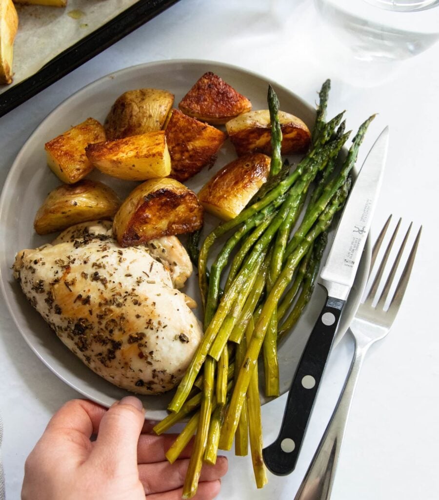 A hand holding a small gray plate with a chicken breast, asparagus, and potatoes.