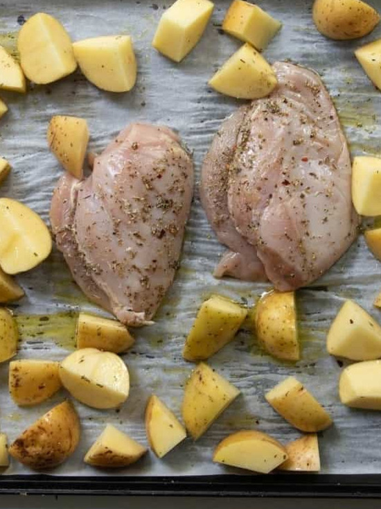 Two raw, seasoned chicken breasts and chopped potatoes sit on a parchment-lined baking sheet—classic sheet pan chicken and potatoes, ready to be cooked.