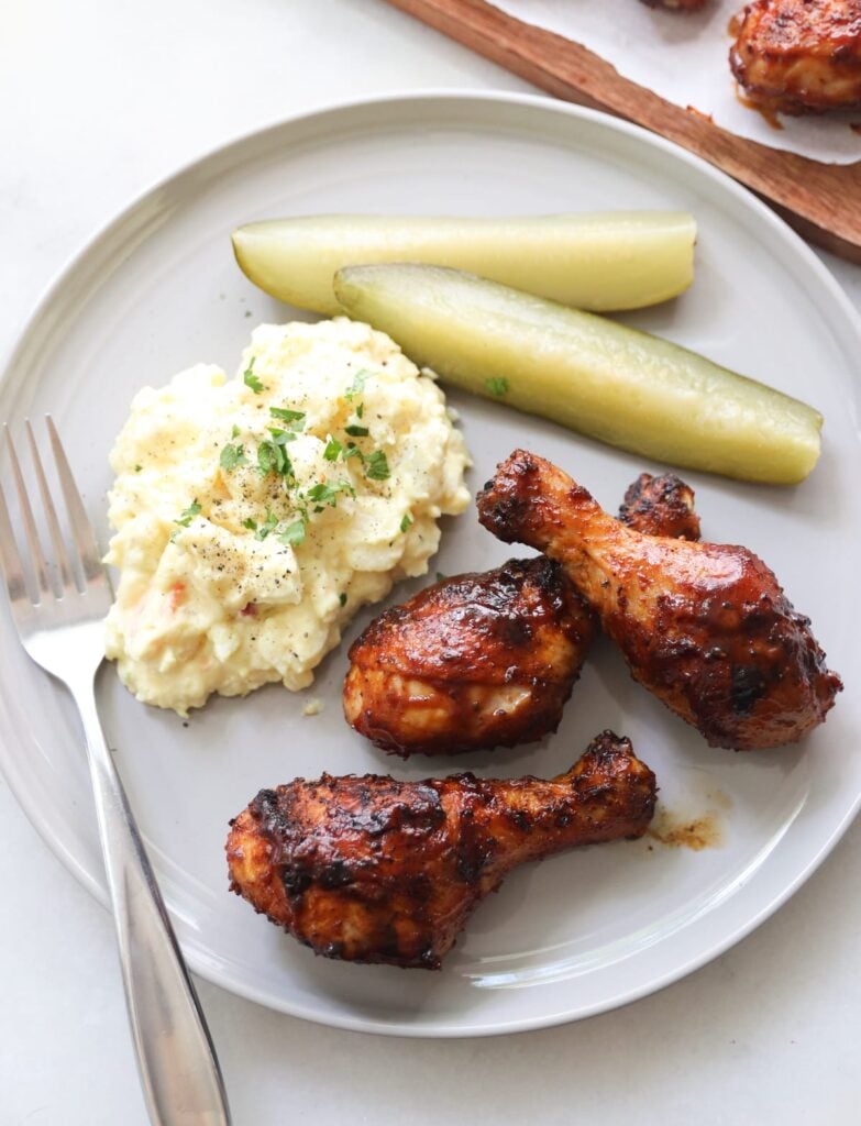 Close up of a gray plate with three bbq chicken drumsticks, potato salad, and pickles.