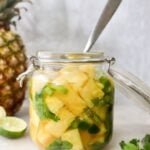 A glass jar brimming with pickled pineapple, cilantro, and sliced green peppers, accompanied by a spoon. Nearby, a fresh pineapple lies beside lime and sprigs of cilantro leaves.