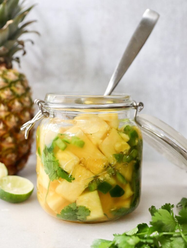 A glass jar brimming with pickled pineapple, cilantro, and sliced green peppers, accompanied by a spoon. Nearby, a fresh pineapple lies beside lime and sprigs of cilantro leaves.