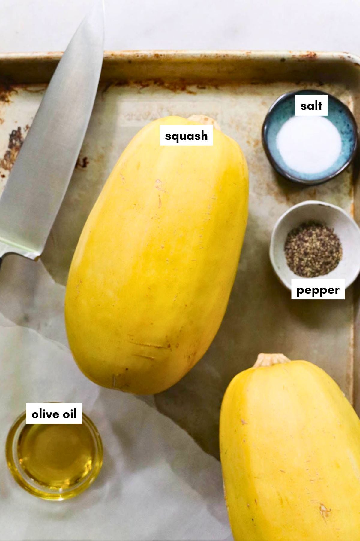 A baking sheet with two winter squashes, a large chef's knife, a small bowl of salt, a small bowl of pepper, and a small bowl of olive oil.