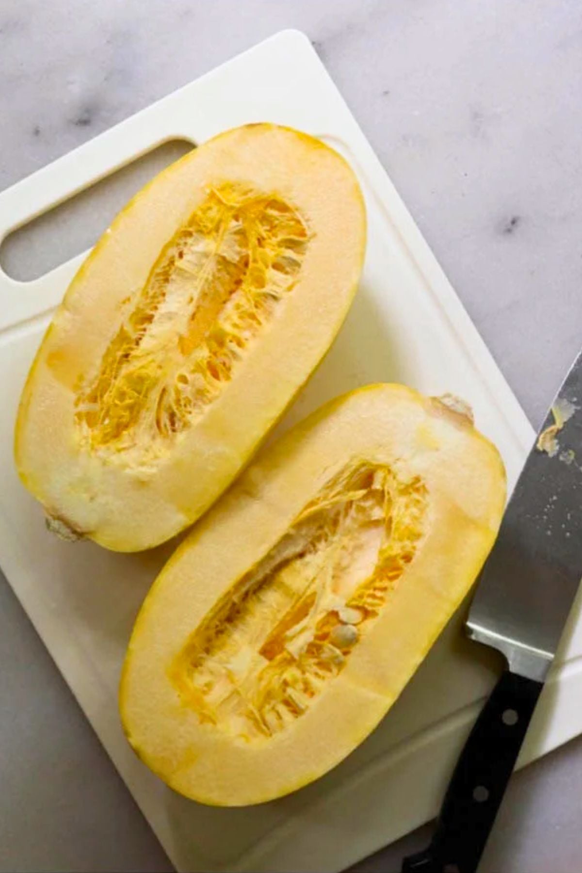 An uncooked squash cut in half lengthwise, placed on a white cutting board next to a knife.