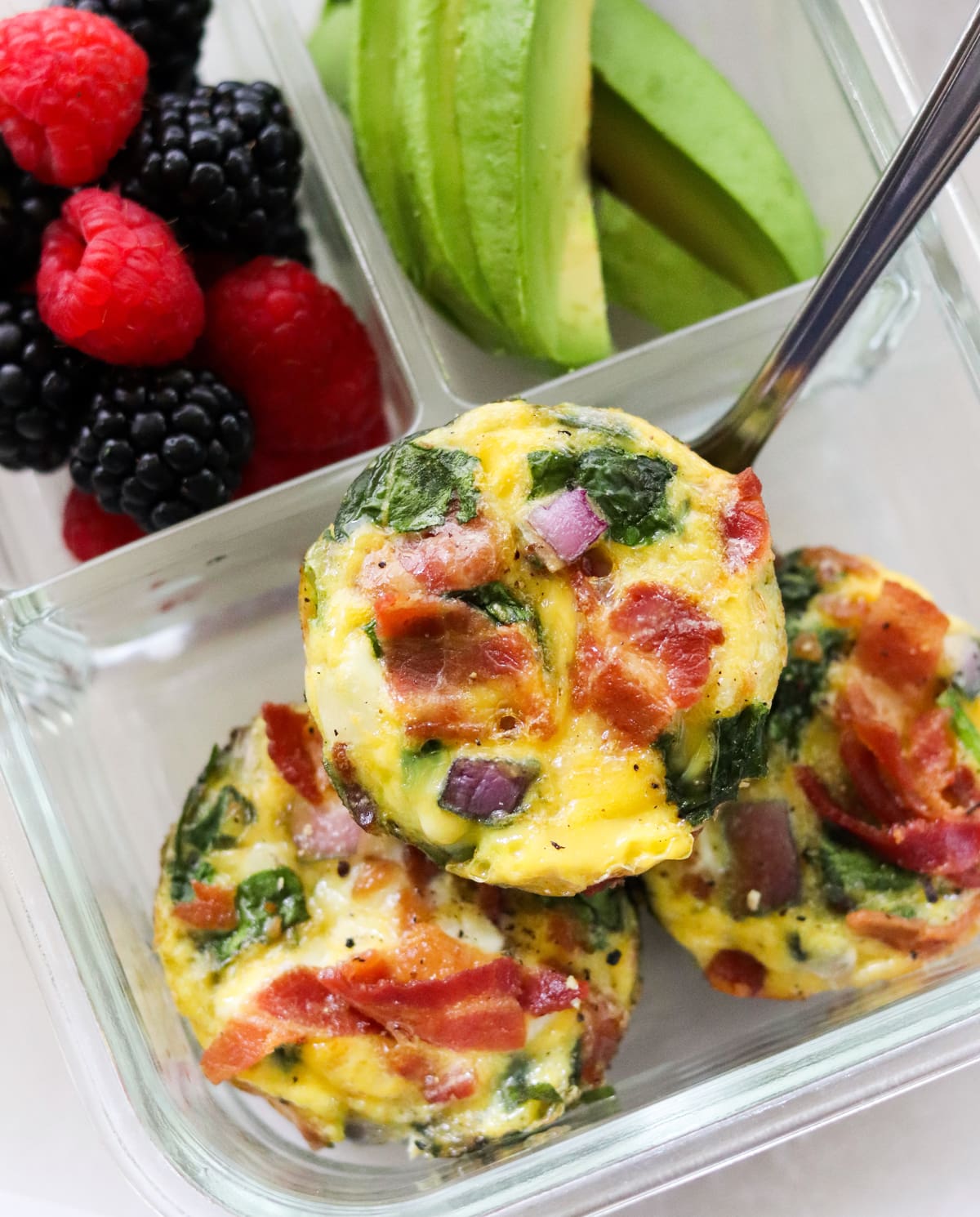 Close up of three bacon and spinach egg muffins in a glass meal prep container with berries and avocado. 