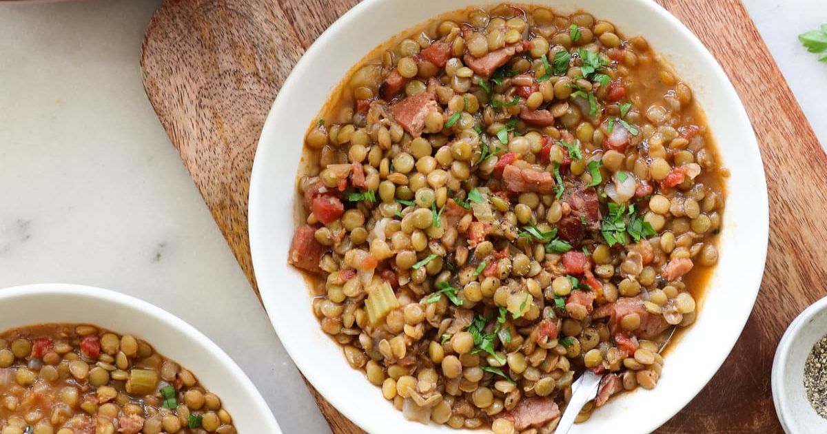 A bowl of ham and lentil soup with diced sausage and herbs on a wooden cutting board.