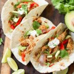 Close up of three plated chicken fajitas topped with cilantro and sour cream on a wooden board.