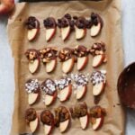 Chocolate dipped apple slices topped with raisins, nuts, or sea salt are arranged in rows on a parchment-lined baking tray.