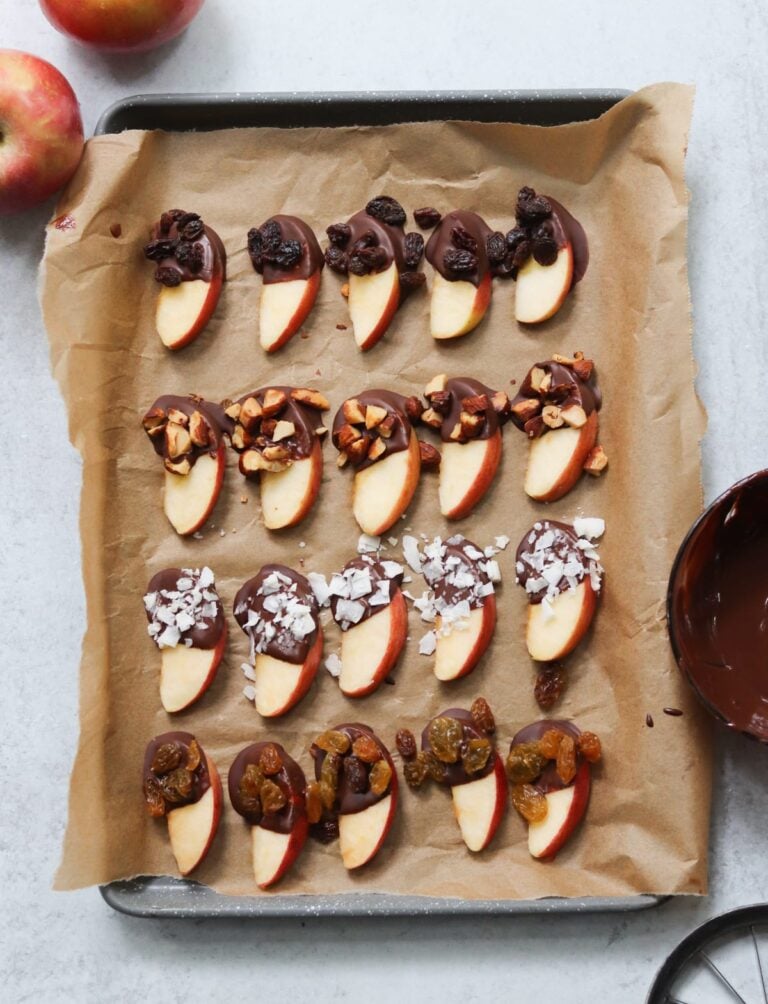 Chocolate dipped apple slices topped with raisins, nuts, or sea salt are arranged in rows on a parchment-lined baking tray.