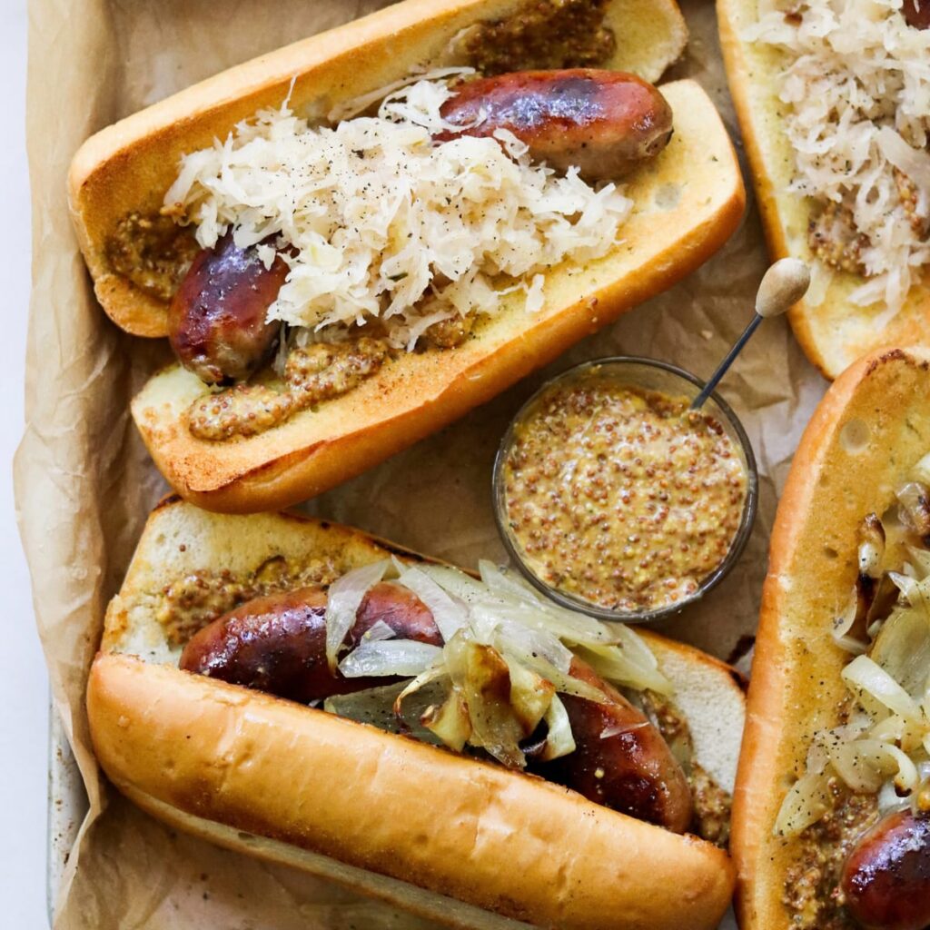 Four brats in toasted buns with sauerkraut and mustard on a baking sheet.