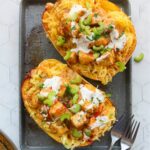 Two stuffed spaghetti squash halves topped with diced chicken, sauce, sliced celery, and chopped green onions on a gray tray.