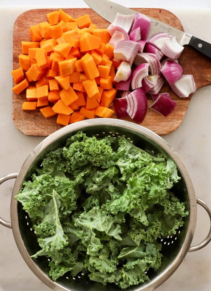 Overhead image of kale leaves in a colander next to chopped butternut squash and red onion slices.