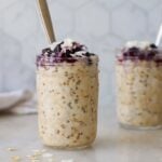 Two mason jars filled with overnight oatmeal that are topped with blueberries and coconut flakes. One of the jars has a spoon in it.