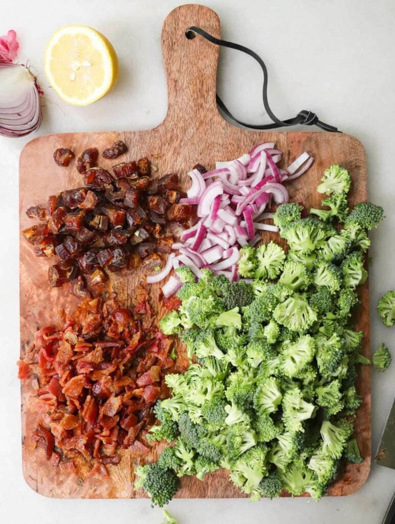 Overhead view of chopped broccoli, chopped cooked bacon, slices of red onion, and chopped dates on a wooden cutting board.