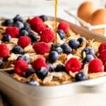 A delicious overnight French toast casserole fills the baking dish, layered with toast, raspberries, blueberries, and sliced almonds, while syrup is being drizzled generously over the top. Eggs and a bowl of berries sit invitingly in the background.