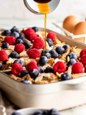 A delicious overnight French toast casserole fills the baking dish, layered with toast, raspberries, blueberries, and sliced almonds, while syrup is being drizzled generously over the top. Eggs and a bowl of berries sit invitingly in the background.