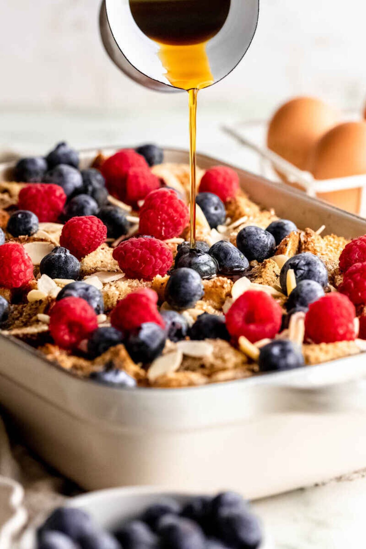A delicious overnight French toast casserole fills the baking dish, layered with toast, raspberries, blueberries, and sliced almonds, while syrup is being drizzled generously over the top. Eggs and a bowl of berries sit invitingly in the background.