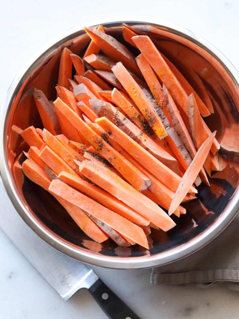 Raw sweet potato fries seasoned with salt and pepper in a metal bowl.