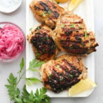 Four grilled pieces of Greek Yogurt marinated chicken on a plate with lemon, parsley, and pickled onions.