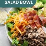 Salad bowl with lettuce, seasoned ground beef, and shredded cheese is perfectly complemented by bacon bits, pickle slices, and sesame seeds. Topped with a creamy dressing reminiscent of a pork chop marinade, it offers a delightful twist. A small side bowl of sesame seeds completes the dish.