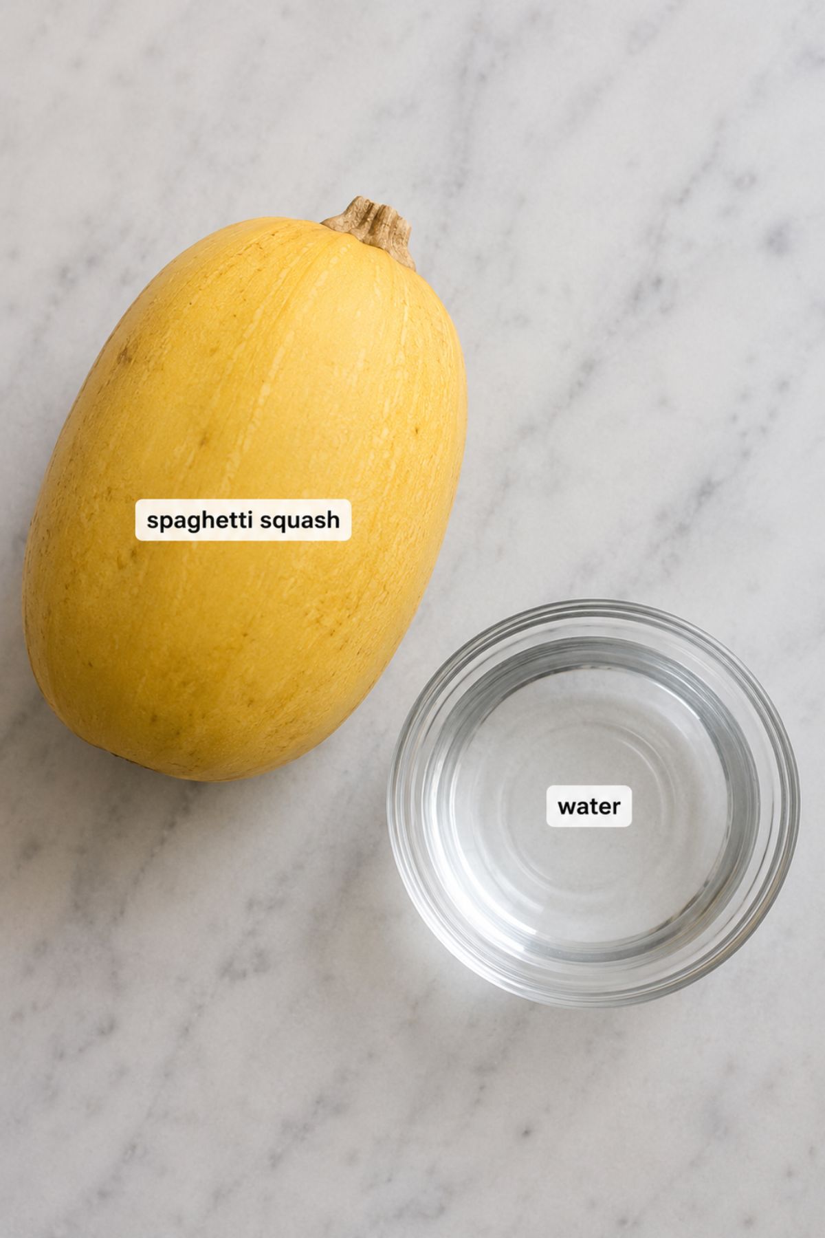 A whole spaghetti squash and a glass bowl of water are placed side by side on a light-colored marble surface.