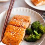 Grilled salmon fillet with sesame seeds and salmon teriyaki marinade served with broccoli on a ceramic plate, accompanied by chopsticks.