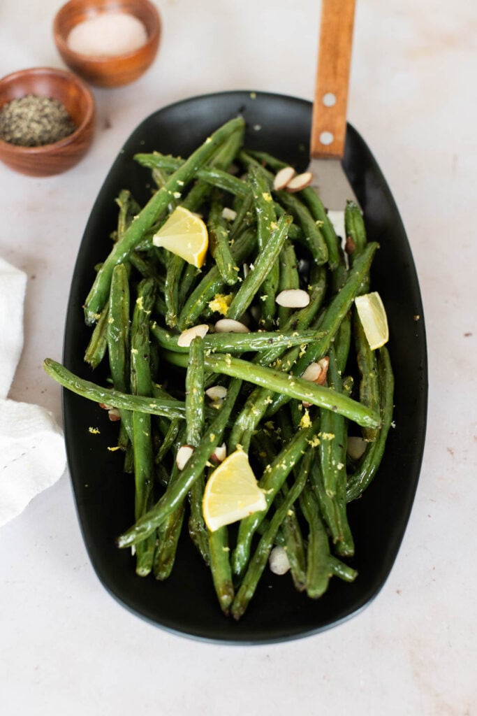 Green beans on a black plate with lemon zest and lemon wedges.