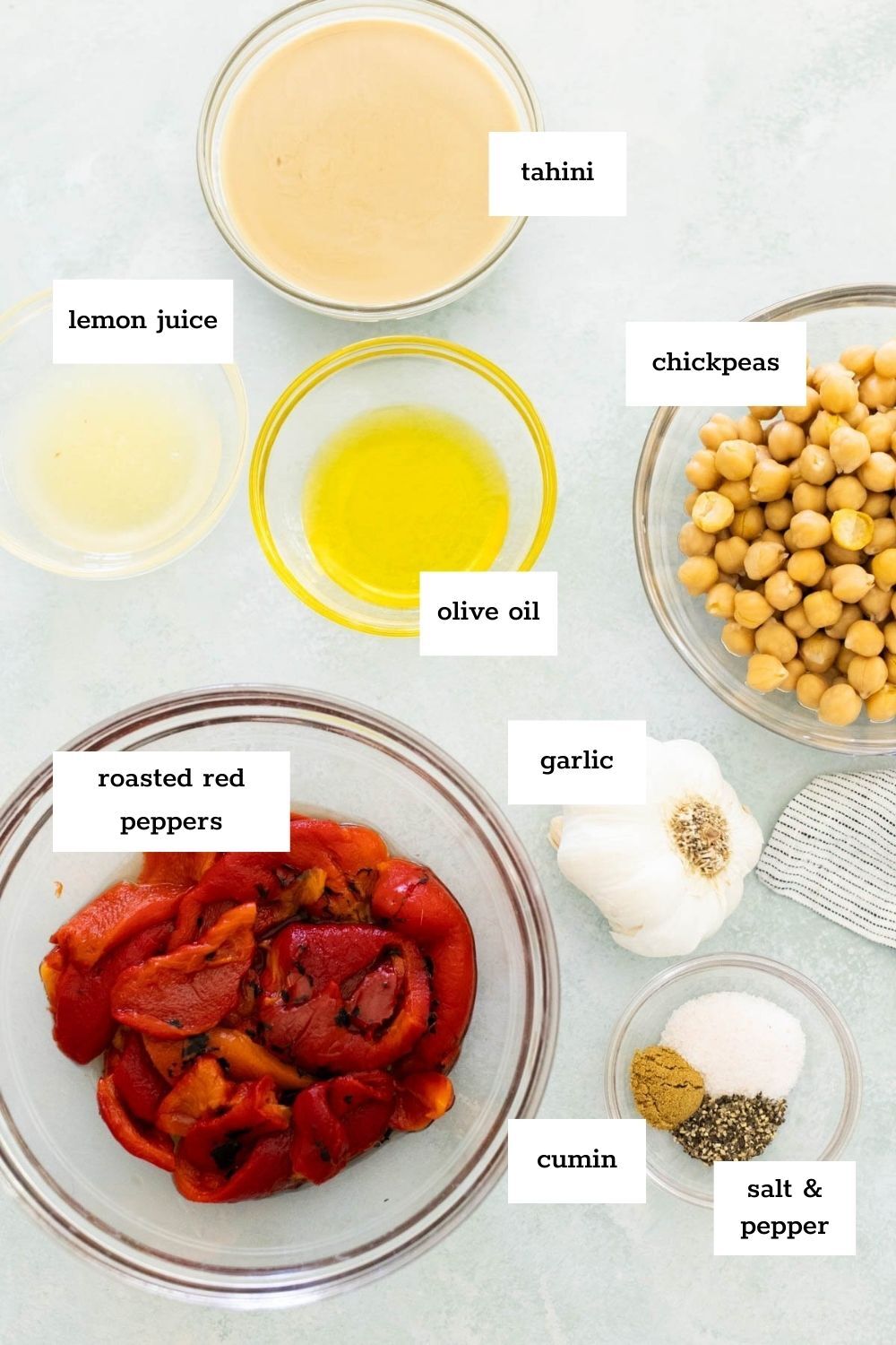 Labeled bowls of ingredients on a light surface: tahini, chickpeas, roasted red peppers, lemon juice, olive oil, garlic, cumin, salt, and pepper.
