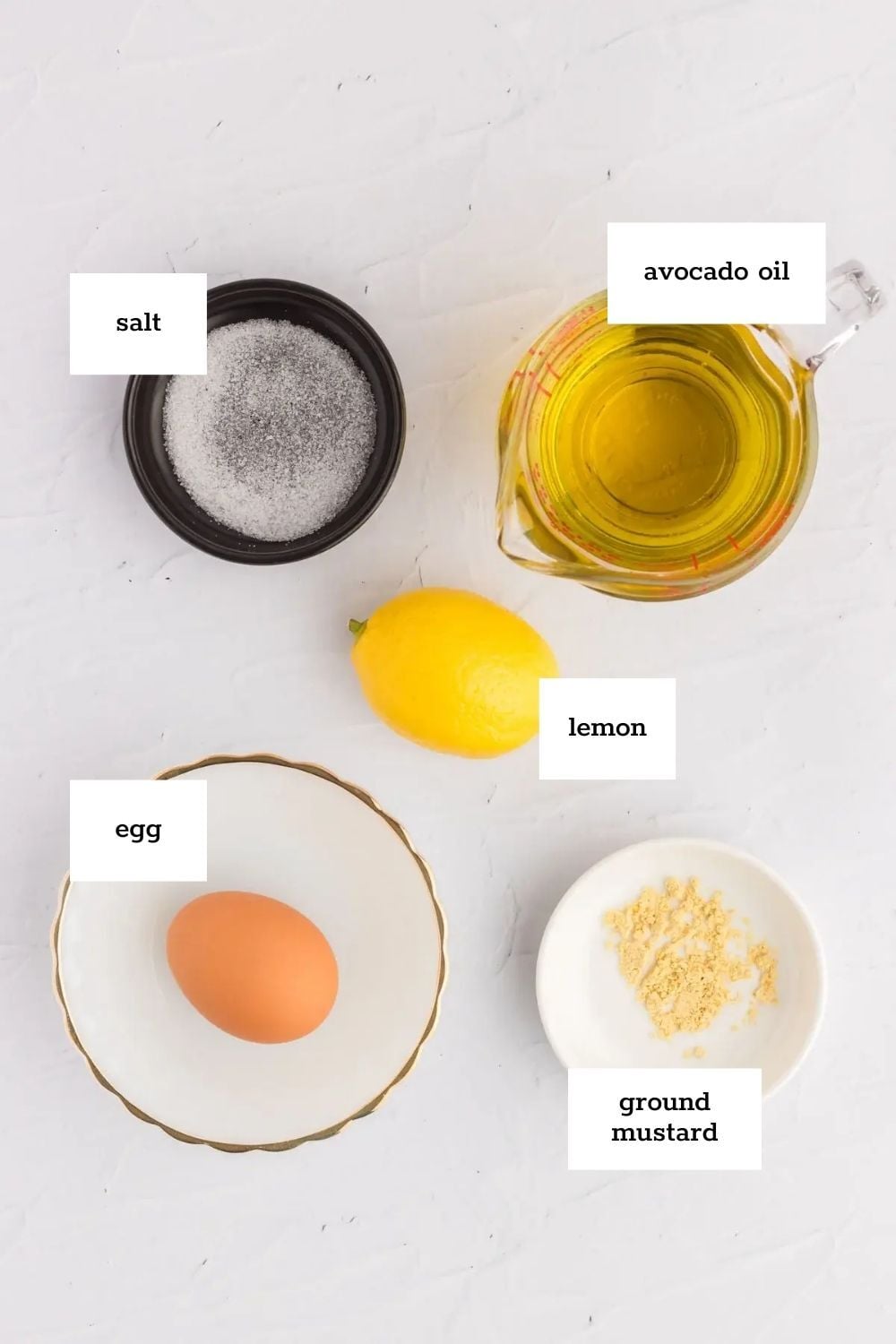 Top-down view of labeled ingredients for avocado mayo on a white surface: salt, avocado oil, lemon, egg, and ground mustard displayed in separate dishes.