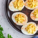 Deviled eggs sprinkled with paprika on a dark plate, garnished with parsley, on a speckled grey surface.
