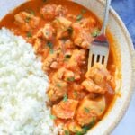 A bowl of butter chicken next to jasmine rice, garnished with herbs, with a fork in the bowl.