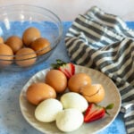 Hard-boiled eggs and fresh strawberries on a plate with a bowl of whole eggs and a striped cloth in the background.