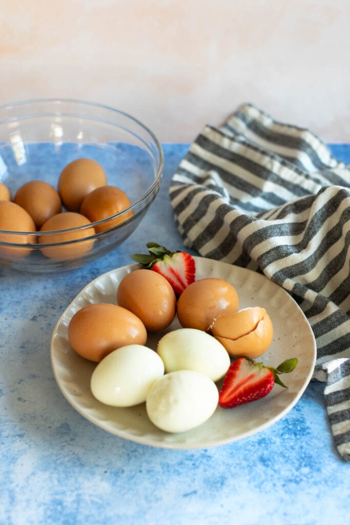 Hard-boiled eggs and fresh strawberries on a plate with a bowl of whole eggs and a striped cloth in the background.