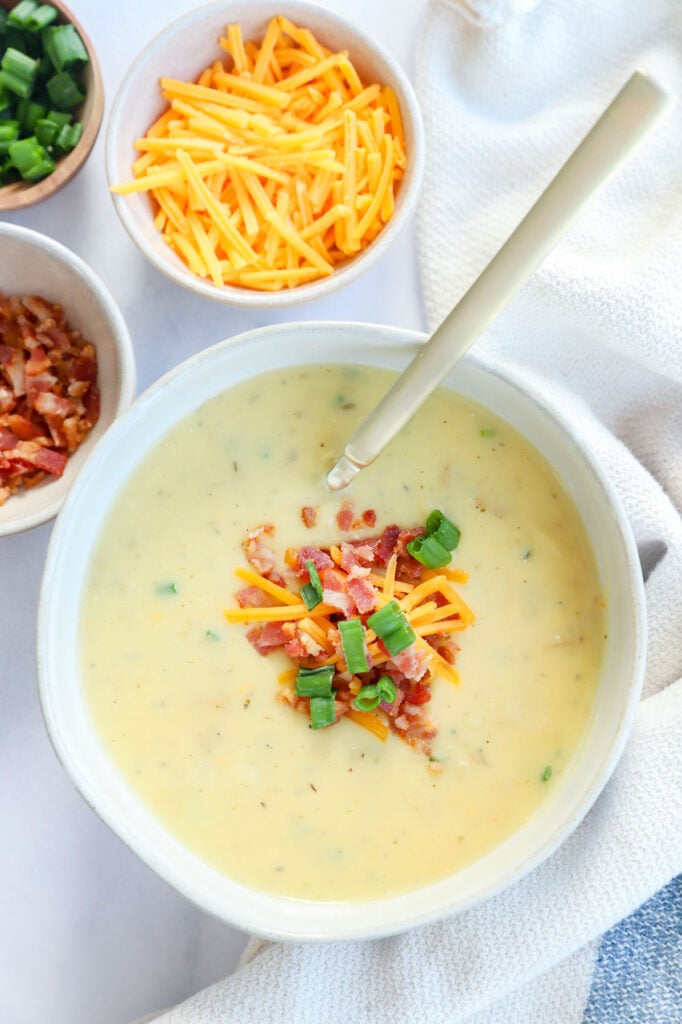 Spoon in a bowl of cheesy potato soup garnished with bacon bits, shredded cheese and chives.