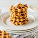 A stack of keto waffles with a pat of butter and syrup being poured on top, served on a white plate.