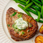 A cooked steak topped with herb butter, accompanied by green beans and roasted potatoes on a white plate.