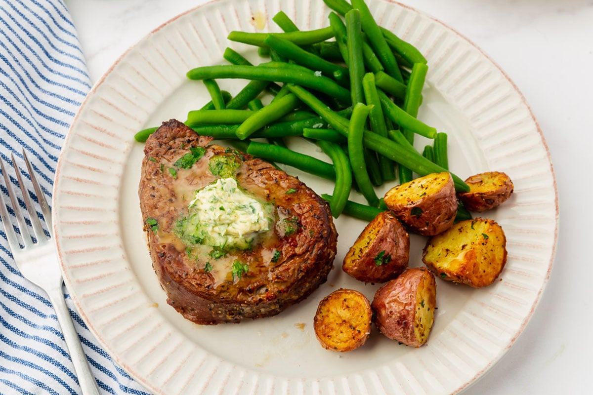 A plate with a cooked steak topped with herb butter, green beans, and roasted potato halves. A striped napkin and fork are placed beside the plate.