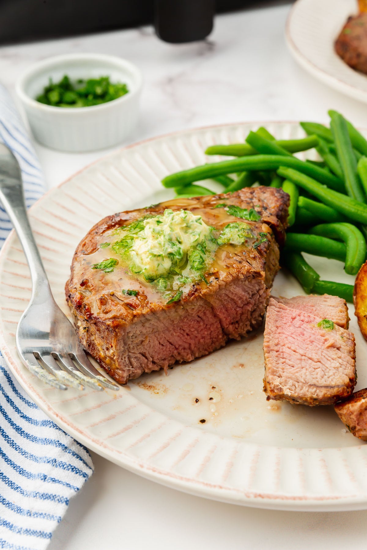 Medium-rare filet mignon steak topped with herb butter, a portion of green beans, and roasted potatoes. A fork rests on the plate.