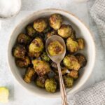 A bowl of roasted Brussels sprouts with a wooden spoon, accompanied by a small bowl of salt, a pitcher of sauce, and a cloth napkin.