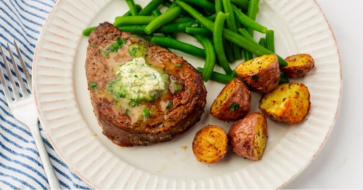 A plate with a cooked steak topped with herb butter, green beans, and roasted potato halves. A striped napkin and fork are placed beside the plate.