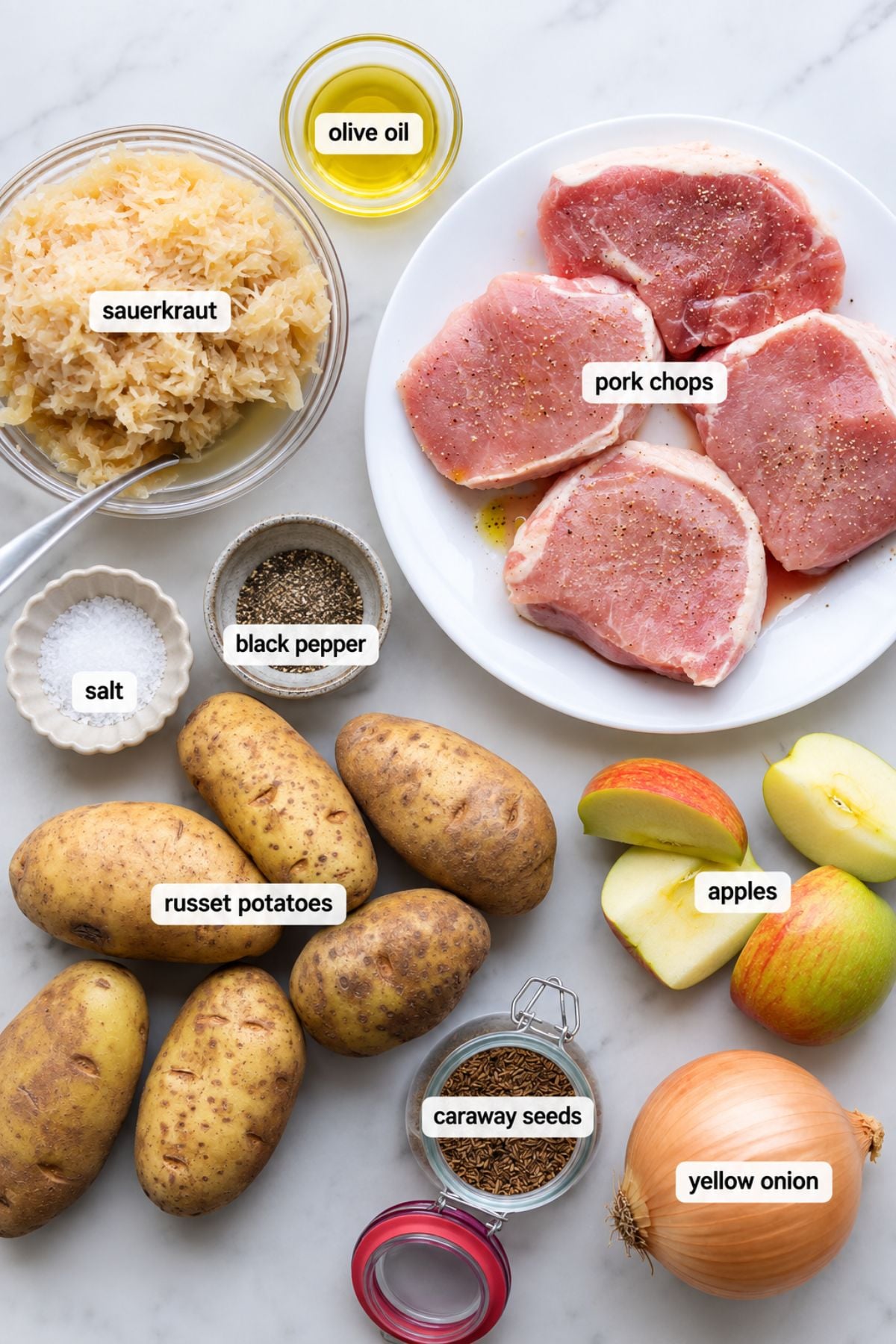 Raw pork chops, sauerkraut, russet potatoes, apples, yellow onion, olive oil, salt, black pepper, and caraway seeds—everything you need for classic German Pork Chops with pork and sauerkraut—are arranged on a countertop in labeled containers.