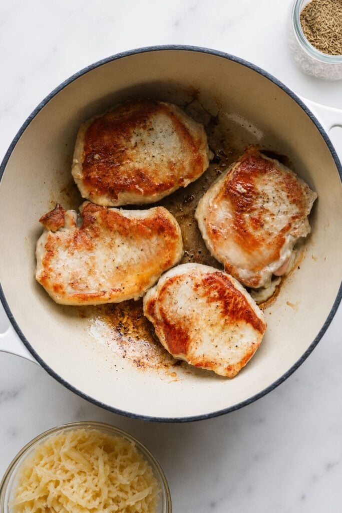Four browned German Pork Chops cook in a white Dutch oven on a marble surface, with bowls of grated cheese and spices nearby, perfect for pairing with tangy sauerkraut.