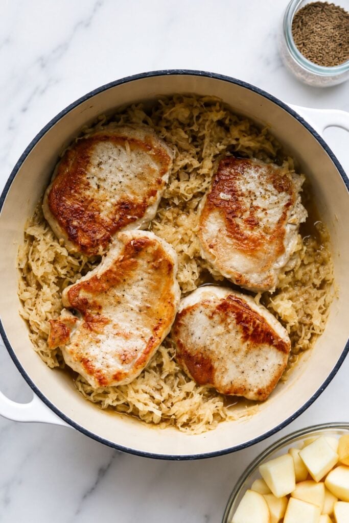 Four browned German Pork Chops rest on a bed of sauerkraut in a round white Dutch oven, seen from above. A small bowl of caraway seeds and a bowl of chopped apples are nearby—perfect accompaniments for this classic Pork and Sauerkraut dish.