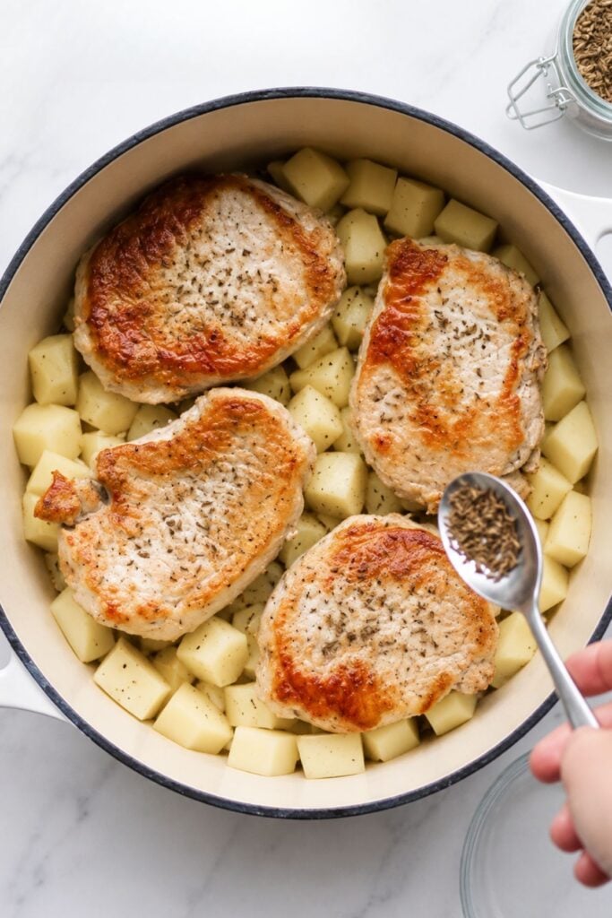 Four browned German Pork Chops rest on a bed of cubed potatoes in a pot, while a hand delicately sprinkles dried herbs from a spoon over the dish.