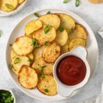 A plate of air fried potato slices garnished with green onions is served with a small container of ketchup. Surrounding items include salt, a whole potato, and a small bowl of chopped green onions.