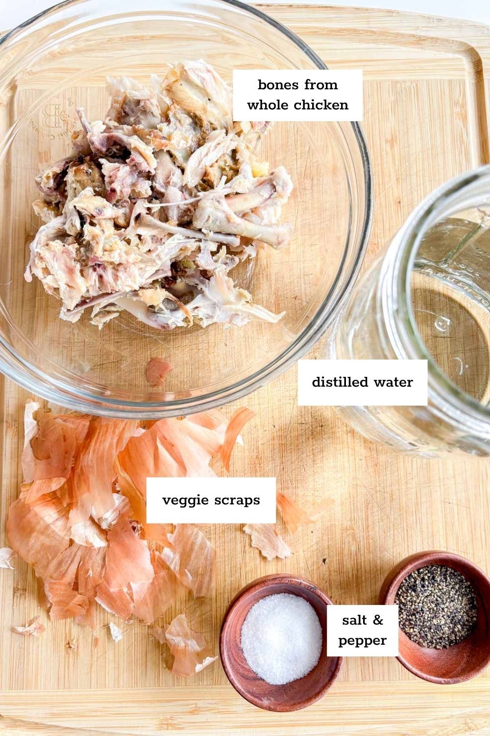 Chicken bones, veggie scraps, salt, pepper, and a jar of distilled water are arranged on a wooden cutting board and in a glass bowl—each labeled for making instant pot chicken broth.