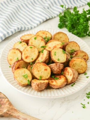 A white plate with sliced, roasted ranch potatoes garnished with chopped parsley. A wooden spoon, striped cloth, and a bunch of parsley are in the background.