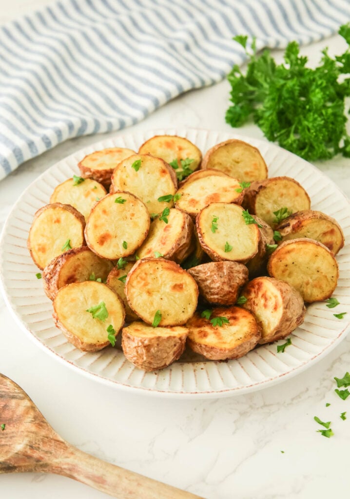 A white plate with sliced, roasted ranch potatoes garnished with chopped parsley. A wooden spoon, striped cloth, and a bunch of parsley are in the background.
