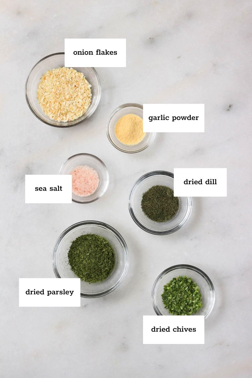 Six small glass bowls on a white surface, each containing a different dried ingredient: onion flakes, garlic powder, sea salt, dried dill, dried parsley, and dried chives.