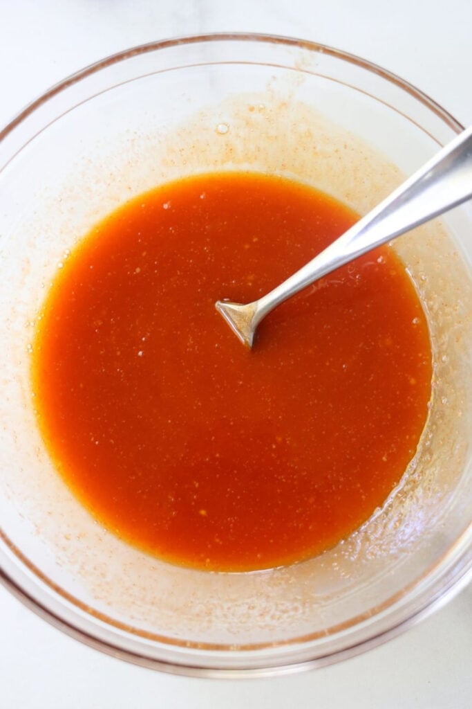 A glass bowl filled with vibrant homemade buffalo sauce, its orange-red liquid gleaming as a metal spoon rests inside.